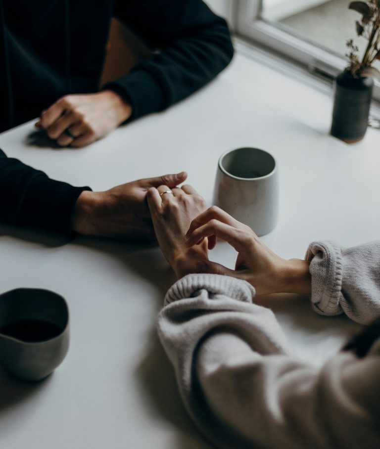 Zwei Personen halten Hände über einem Tisch auf dem zwei Tassen und eine kleine Pflanze stehen