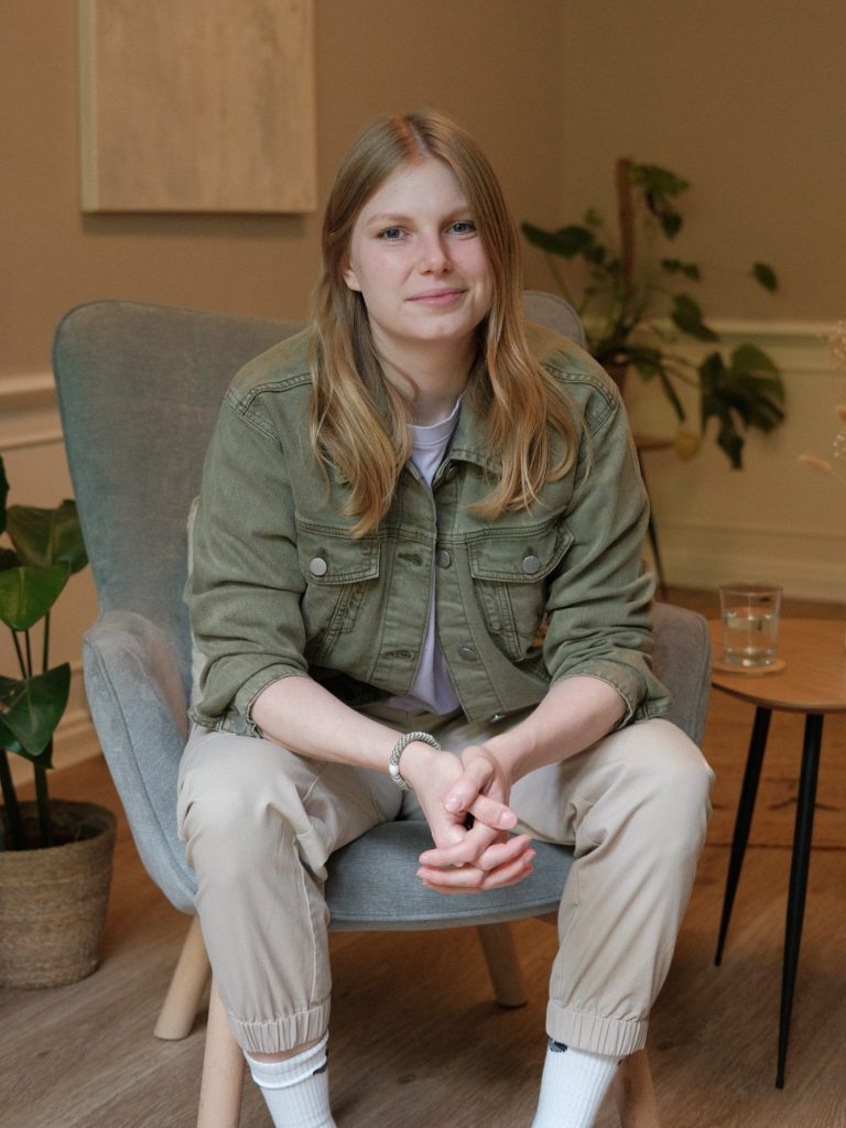 Das Bild zeigt die Beraterin Janine Kröger im Praxisraum. Eine junge Frau mit langen Haaren sitzt in einem Sessel, umgeben von Pflanzen. 