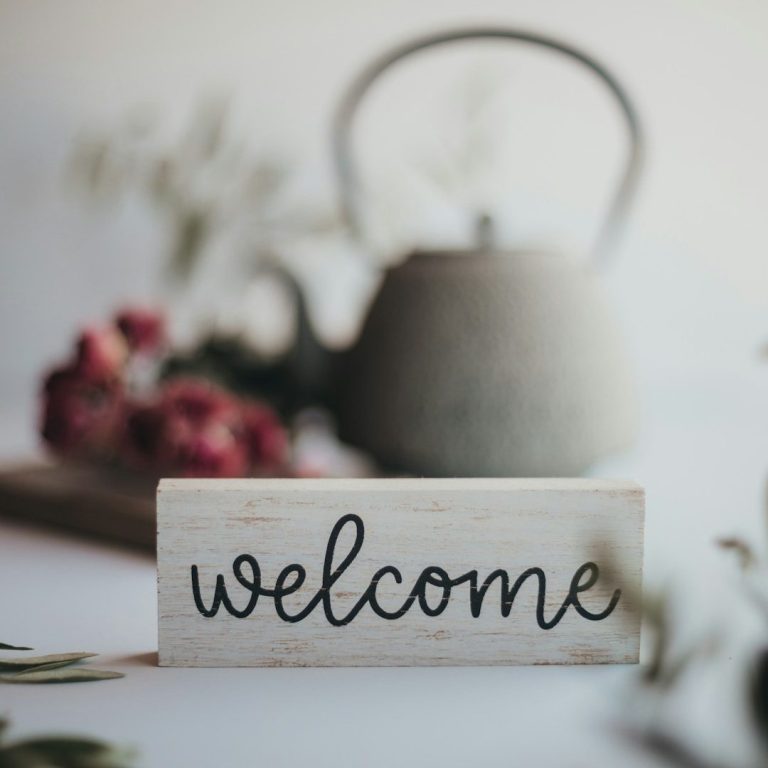 Willkommensschild mit geschwungener Schrift, Blüten und einer Teekanne im Hintergrund.