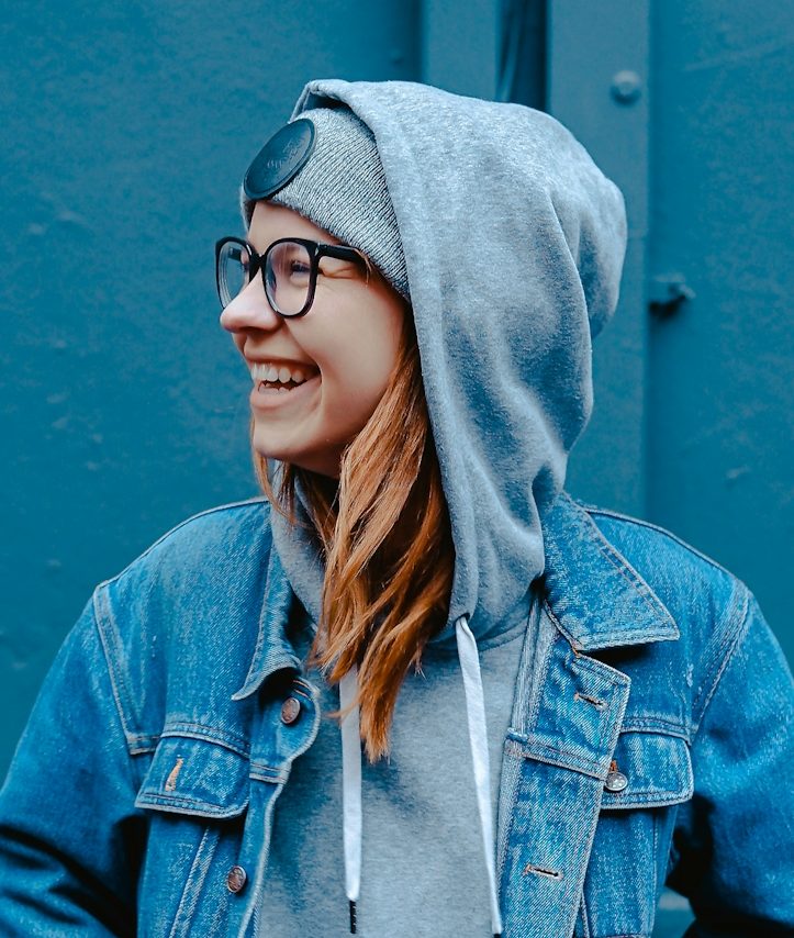 Junge Frau mit Brille, Hoodie und Jeansjacke, lächelt vor einer blauen Wand.