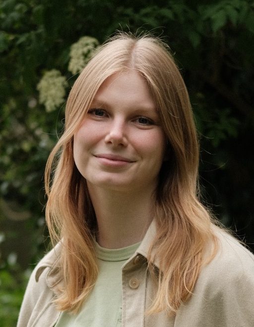 Das Bild zeigt ein Portrait der Beraterin Janine Kröger, eine junge Frau mit langen, blonden Haaren, lächelnd vor grünen Pflanzen.